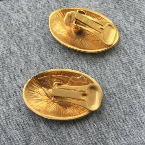 May include: A pair of gold-coloured clip-on earrings. Each earring is oval-shaped with a textured surface and a clip-on mechanism. The earrings are displayed on a grey surface.