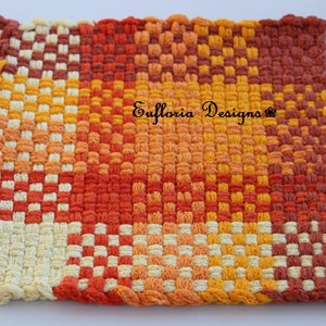 May include: A woven pot holder with a checkered pattern in shades of orange, yellow, and white. The pot holder has a loop for hanging and the text "Eufloria Designs" is woven into the center.