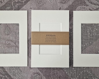 Op de afbeelding: Drie gebroken witte, rechthoekige passe-partouts met centrale uitsparingen. Een bruine papieren band zit om het middelste passe-partout, bedrukt met de tekst: ACEO Mounts, Textured Off-White, Conservation Quality, Acid-free & Archival, To fit a 4" x 6" frame.