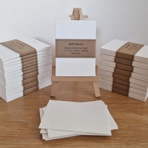May include: Stacks of white oil painting paper blanks, each bound with a brown paper band. The text "ACED Blanks" is visible on the band. A small wooden easel holds a set of the paper blanks. A few loose sheets are in the foreground.