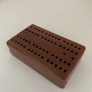 May include: A rectangular dark brown wooden cribbage board featuring a series of drilled holes arranged in parallel rows across the top surface. The board has a smooth finish and a solid construction with visible wood grain patterns throughout.