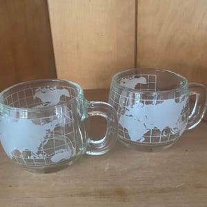 May include: Two clear glass mugs featuring a white etched world map design with a grid of latitude and longitude lines. Each mug has a rounded bowl shape and a sturdy glass handle on the side, showcasing a detailed global cartographic pattern.