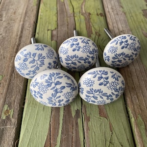 May include: Five white ceramic cabinet knobs with a blue floral pattern. Each knob has a silver metal screw. The knobs are arranged on a weathered, green-painted wooden surface.