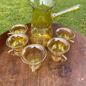 May include: A set of six amber glass cups and a matching pitcher with a handle. The pitcher has a label that reads "Otto Kroglass Handgjort Glas."
