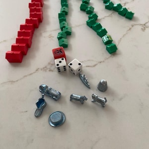 May include: A collection of Monopoly game pieces, including red and green houses, dice, and metal tokens like a car, shoe, dog, and top hat. The pieces are arranged on a light-colored surface.