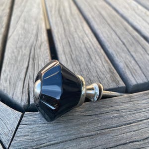 May include: A faceted black cabinet knob with a glossy finish and a silver-toned metal base. The knob features a central silver-colored circular accent on its front face and includes a threaded metal screw extending from the back for installation.