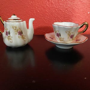 May include: A white tea set with gold trim and floral designs. The set includes a teapot and a cup and saucer.