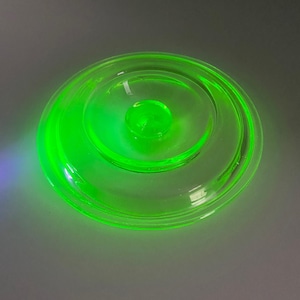 May include: A clear, round glass lid with a bright neon green glow. The lid has a central knob and concentric rings, creating a layered effect. The glass appears to be vintage or antique, with a distinctive color.