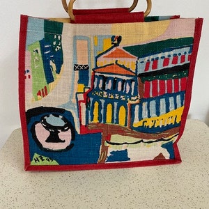 May include: A colorful tote bag with a red border and bamboo handles. The bag features a graphic design of a building with abstract shapes and lines in shades of blue, green, yellow, red, and orange.