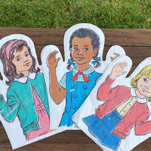 May include: Three vintage oven mitts with illustrations of young girls. Each mitt features a different girl in a colorful outfit. The mitts are white with the girls' names printed at the bottom. The background is a wooden surface with green grass.