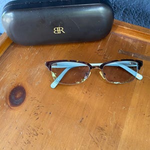 May include: Eyeglasses with a brown tortoiseshell frame and light blue arms, resting on a wooden surface. A black eyeglasses case with a gold "BR" logo is also present.