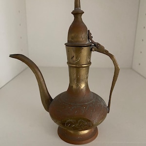 May include: A vintage brass teapot with a long spout and ornate handle. The teapot has a bulbous body with etched floral designs and a tall, pointed lid. The brass has a warm, aged patina, suggesting it is an antique or vintage piece.