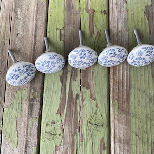 May include: Five oval ceramic cabinet knobs with a white base and blue floral pattern. Each knob has a silver metal screw for installation. The knobs are arranged in a row on a weathered wooden surface with peeling green paint.