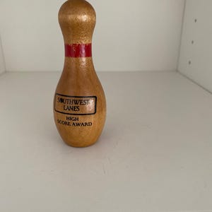 May include: A wooden bowling pin trophy with a smooth, light brown finish. It features a single horizontal red stripe around the neck and black text that reads SOUTHWEST LANES HIGH SCORE AWARD printed on the front of the body.