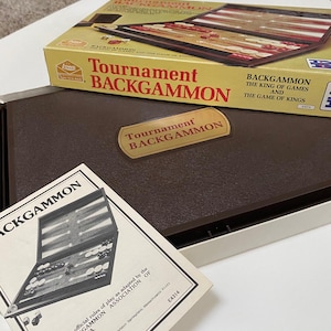 May include: A brown wooden backgammon game set with a brown leatherette case. The box has the text "Tournament Backgammon" and "Backgammon The King of Games and The Game of Kings". The game includes a set of instructions with the text "The official rules of play as adapted by the Backgammon Association of America."