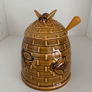 May include: A brown ceramic honey pot with a lid and a small spoon. The pot is shaped like a beehive and has a bee on top of the lid. Two additional bees are on the side of the pot. The honey pot is a decorative kitchen item.
