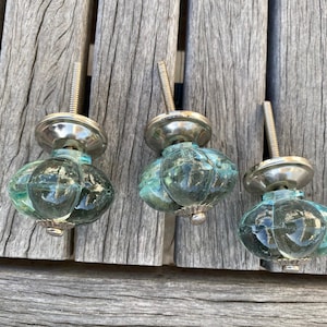 May include: Three cabinet knobs featuring translucent, seafoam green glass bodies with a rounded, ribbed, and faceted design. Each knob includes a polished silver-tone metal base and a threaded metal bolt extending from the back for installation.