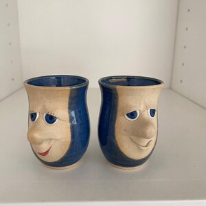 May include: Two ceramic face mugs. Each mug has a tan face with blue eyes, a nose, and a red smiling mouth. A vertical blue stripe runs down the side of each mug, and the inside is also blue.