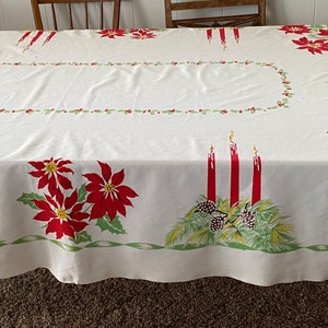 May include: White rectangular tablecloth with a festive Christmas design. The tablecloth features red poinsettias, green holly leaves, and red candles. The border has a green and white ribbon design. The tablecloth is on a wooden table.
