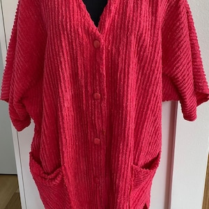 May include: A bright red, textured, button-down cardigan with short sleeves and two front pockets. The garment has a V-neck and is displayed on a black mannequin head. The fabric has a vertical ribbed pattern.