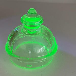 May include: A translucent, vibrant neon green glass lid featuring a rounded dome shape and a central knob handle. The glass exhibits a bright, glowing appearance with subtle vertical ridges along the curved surface and a smooth, circular rim.