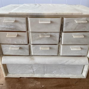 May include: A vintage jewelry box with a marbled, off-white finish. The box features nine small drawers and a larger bottom drawer, all with matching cream-colored handles. The box is made of a sturdy, possibly plastic, material.