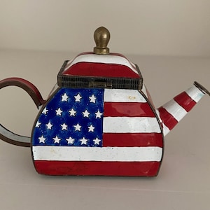 May include: A decorative teapot featuring the American flag design. The teapot has a red, white, and blue color scheme with a brass-colored knob on the lid. The handle and spout are also red and white striped.
