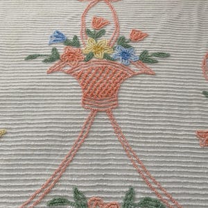 May include: A white fabric with a textured horizontal striped pattern featuring embroidered designs of a peach-colored basket filled with yellow, blue, and peach flowers, accented by green leaves and a peach bow, with additional floral motifs below.