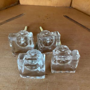 May include: Four clear glass cabinet knobs with a square base and a circular top section. Each knob features a metallic screw extending from the back, designed for attachment to furniture doors or drawers, showcasing a transparent design.
