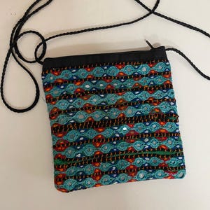 May include: A small, square crossbody bag with a black strap. The bag features a colorful, embroidered design with teal, orange, and black threads, and small reflective accents. The top of the bag is black.