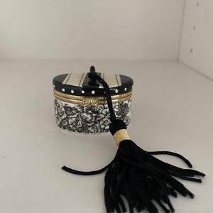 May include: A small, round decorative box with a black and white striped lid and a black tassel. The box features a floral pattern in black and white and is trimmed with gold accents. The tassel is attached to the lid with a black cord.