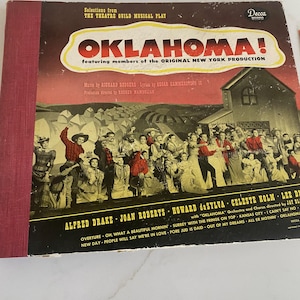 May include: Vintage vinyl record album cover for the musical "Oklahoma!" featuring the original New York production. The cover is predominantly brown and red, with the title in a yellow and white banner. The album features a cast photo.