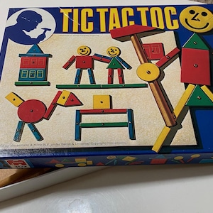 May include: A blue and yellow Tic Tac Toc game box with a smiling face on the front. The box contains a set of colorful wooden pieces that can be used to build different structures. The game is made by Hausemann & Hötte N.V. under Berne & Universal Copyright Conventions.