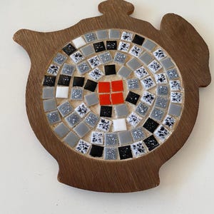 May include: A wooden trivet in the shape of a teapot, featuring a mosaic tile design. The mosaic has a central square of red tiles, surrounded by gray, black, white, and patterned tiles. The trivet is made of dark brown wood.