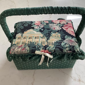May include: A rectangular fabric sewing basket with a green handle and a green base featuring a small white floral pattern. The lid is padded and covered in a dark fabric with a floral print and a central illustration of a large house, finished with a small cream-colored ribbon bow and a ruffled edge.