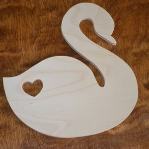 May include: A wooden cutout of a swan with a heart-shaped hole in the body. The swan is light brown and the heart is cut out of the wood.