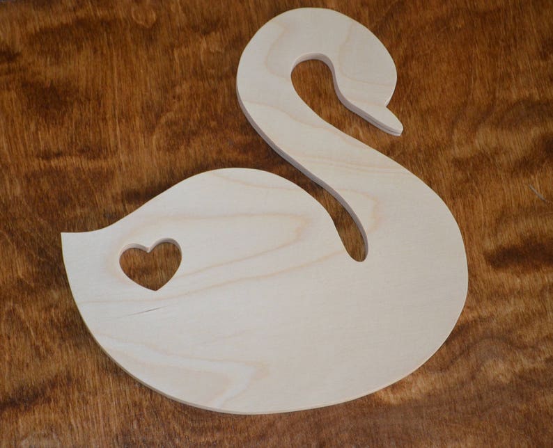 May include: A wooden cutout of a swan with a heart-shaped hole in the body. The swan is light brown and the heart is cut out of the wood.