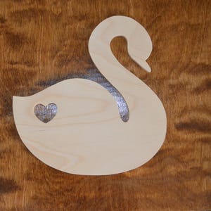 May include: A wooden cutout of a swan with a small heart in the center of the body. The swan is light brown and the heart is a darker brown.