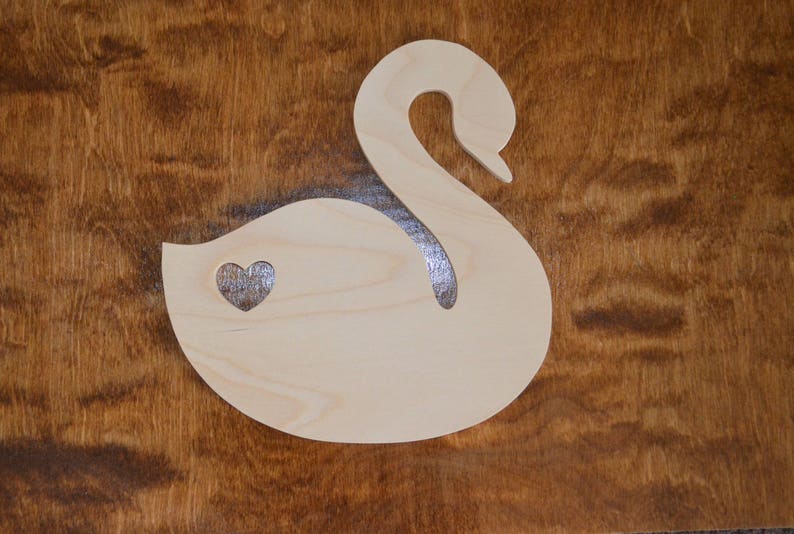 May include: A wooden cutout of a swan with a small heart in the center of the body. The swan is light brown and the heart is a darker brown.