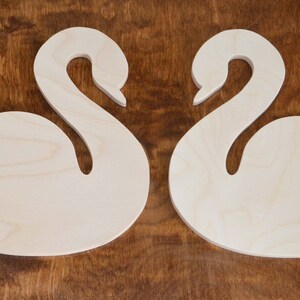 May include: Two unfinished wooden swan shapes with a heart cut out of each. The swans are facing each other.