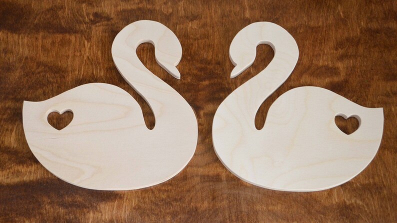 May include: Two unfinished wooden swan shapes with a heart cut out of each. The swans are facing each other.
