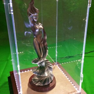 Könnte beinhalten: Eine silberfarbene Skulptur einer Frau in einem fließenden Kleid wird in einer durchsichtigen Acrylvitrine mit einem Holzsockel ausgestellt. Der Sockel trägt den Schriftzug "Acrylicjob".