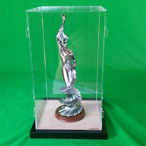 May include: A silver-colored sculpture of a woman in a flowing dress, displayed in a clear acrylic display case with a wooden base.