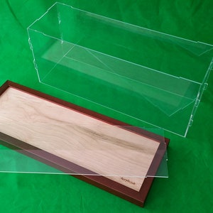 Op de afbeelding: Een transparante acryl vitrine met een houten basis. De basis is gemaakt van licht hout met een bruin frame. De vitrine is open en de basis staat op een groene ondergrond.