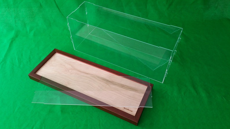 Op de afbeelding: Een transparante acryl vitrine met een houten basis. De basis is gemaakt van licht hout met een bruin frame. De vitrine is open en de basis staat op een groene ondergrond.