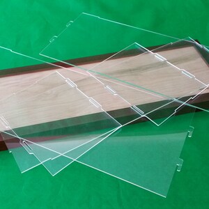 Op de afbeelding: Drie transparante acrylaatplaten met afgeronde hoeken en uitsparingen aan de randen, gestapeld op een houten frame met een lichtbruin houten oppervlak.