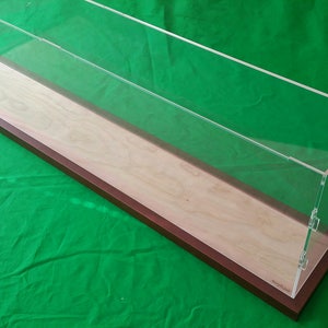 Puede incluir: Una vitrina de acrílico transparente con una base de madera. La base es de madera clara con un borde marrón oscuro. La vitrina es rectangular y tiene una tapa con bisagras.