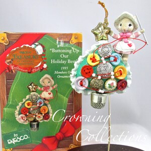 May include: A Christmas tree ornament featuring a small mouse figurine sewing buttons onto a green Christmas tree. The tree is decorated with colorful buttons, beads, and a small pink rose. The ornament is part of the Enesco Treasury of Christmas Collectors Club.