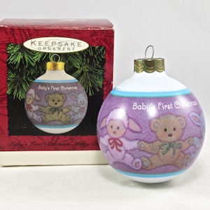 May include: A white glass Christmas ornament with a purple background featuring a teddy bear, a lamb, and a bunny. The ornament has the text "Baby's First Christmas" on it.