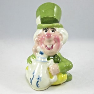 May include: A ceramic figurine of the Mad Hatter from Alice in Wonderland. He is wearing a green hat and a green jacket. He is holding a white teapot with blue and white floral designs.